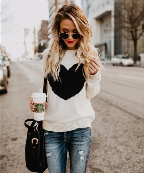 Woman wearing a white sweater with a black heart design, holding a Starbucks cup on a city street.