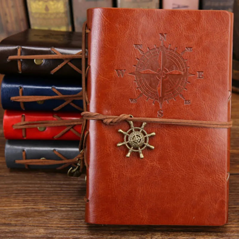Brown leather notebook with compass design and decorative rope, set against a wooden surface with other notebooks.